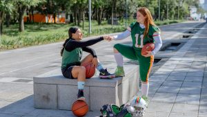 Two female athletes share a fist bump while sitting outdoors, holding sports equipment in Mexico City.